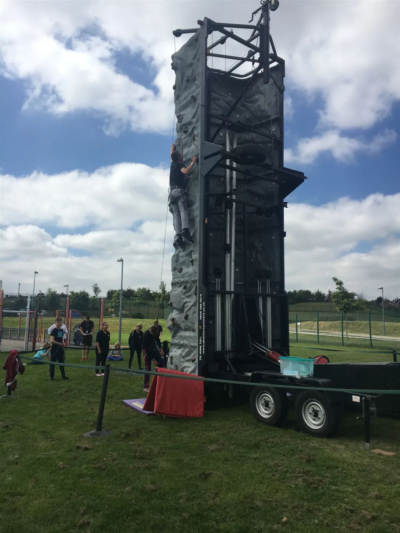 Person climbing a tall mobile climbing wall on a grassy field with spectators nearby