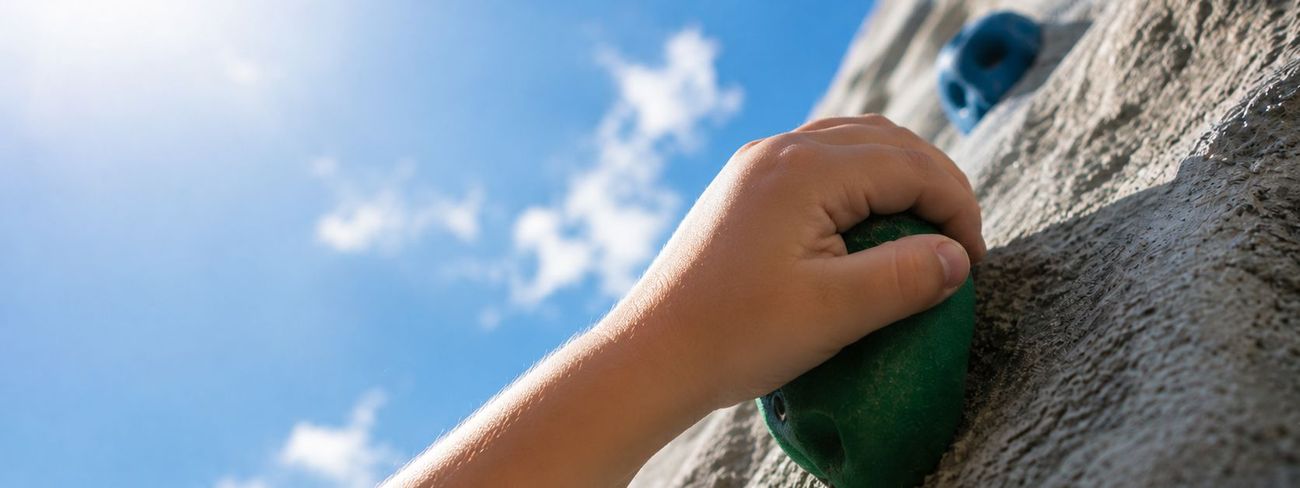 Close-up of a child's hand gripping a green climbing hold against a blue sky