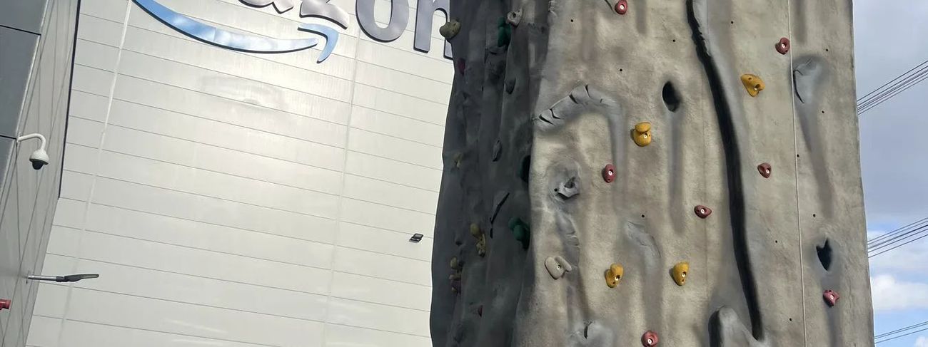 Two climbers ascend a tall mobile climbing wall outside an Amazon building while an instructor watches below
