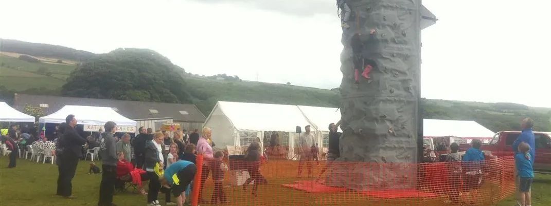 Children and adults climb a mobile climbing wall at an outdoor event with spectators and tents nearby