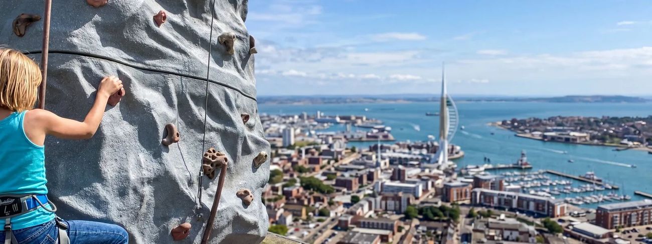 A climber on our mobile climbing wall high above Portsmouth harbour, with the Spinnaker Tower and marina behind